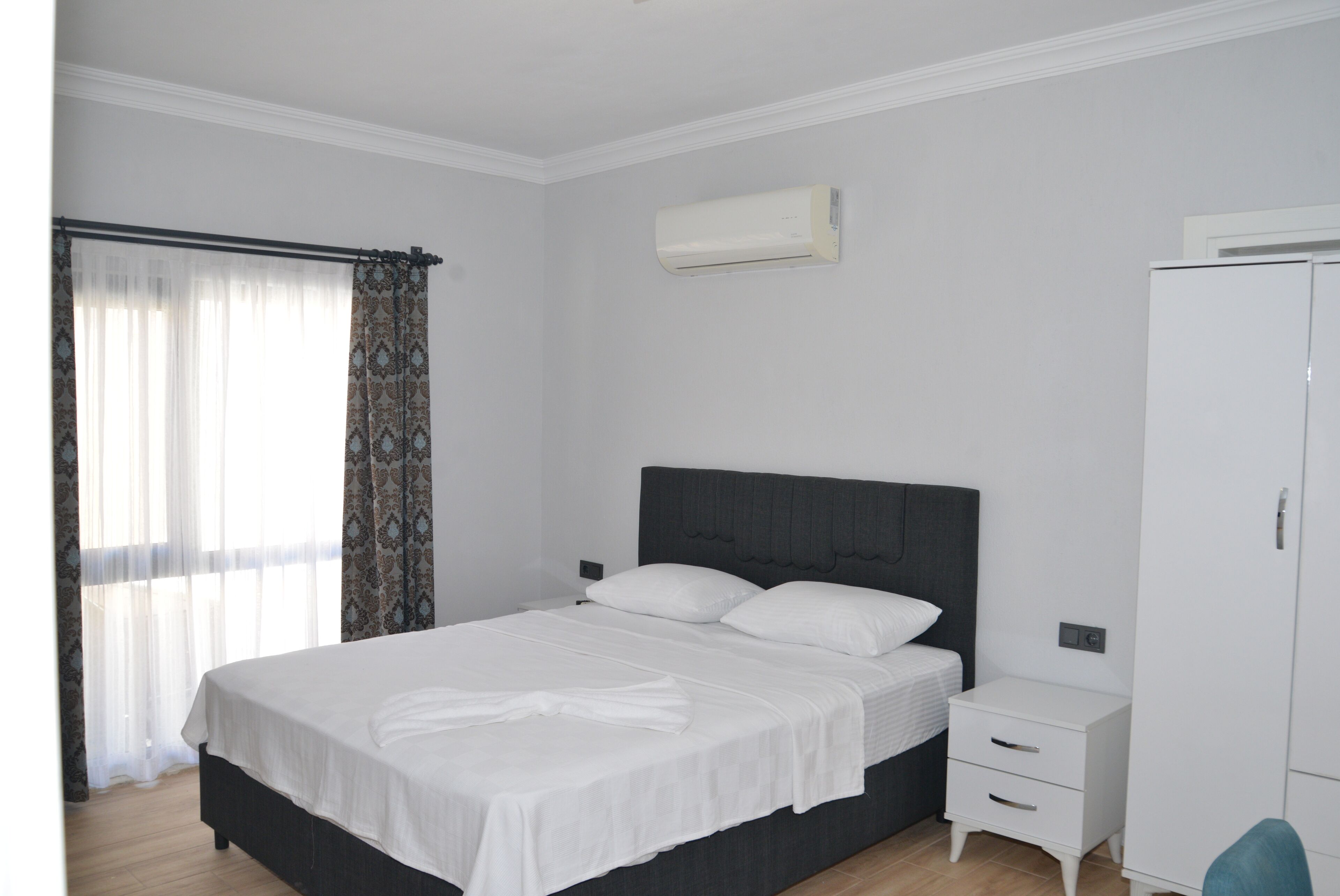 Un dormitorio con cama, mesita de noche, armario y ventana con cortinas.