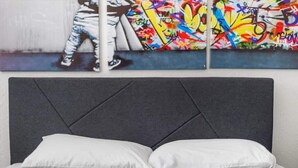 Una cama con cabecera tapizada en gris, almohadas blancas y una pared con un mural estilo graffiti.