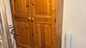 Une armoire en bois à deux portes et une étagère inférieure, placée contre un mur où se trouvent un radiateur et un câble.