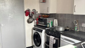 Une cuisine compacte équipée d’un lave-linge, d’un four à micro-ondes et d’un four traditionnel. Il y a une horloge au mur et une étagère contenant des ustensiles de cuisine.