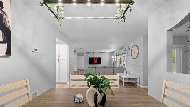 Een modern interieur met een houten eettafel, een potplant, een glazen salontafel, een witte bank en een televisie waarop het Netflix-logo te zien is.