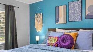 Een slaapkamer met een bed, een macramé wanddecoratie en twee ingelijste kunstwerken.