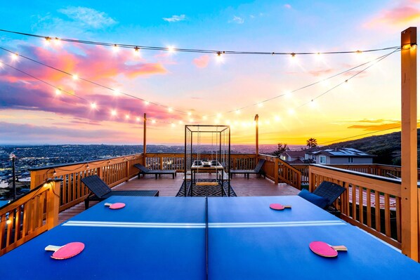 A deck with ping pong tables, a swing set, and string lights against a sunset view.