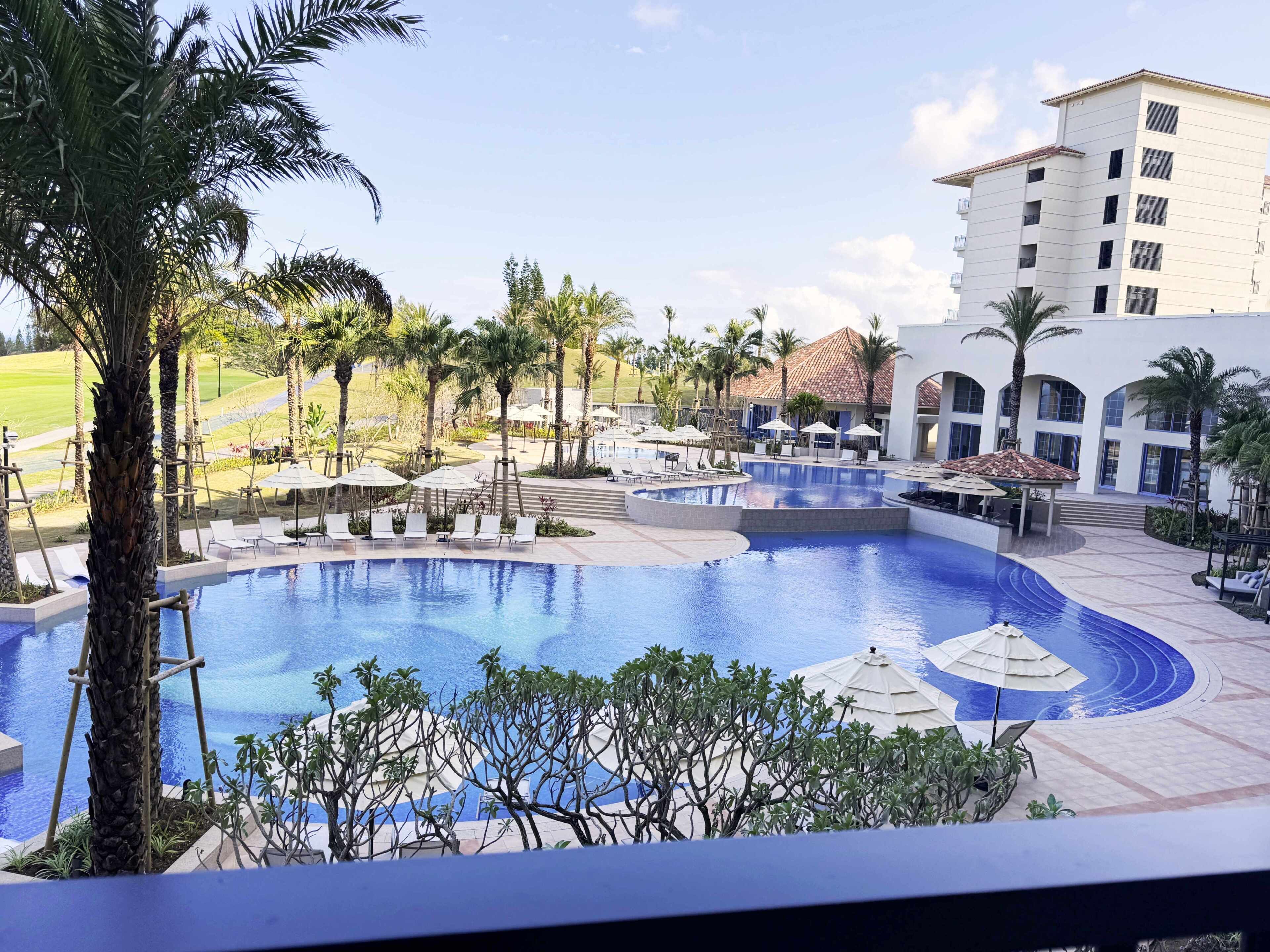 A resort pool with lounge chairs, palm trees, and a building with a tiled roof.
