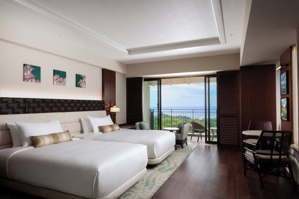 A hotel room with two beds, a balcony, a dining area, and artwork on the wall.