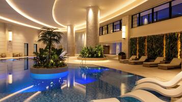 An indoor swimming pool with lounge chairs, a palm tree, and a high ceiling with recessed lighting.