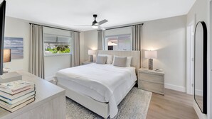 A modern bedroom with a large bed, bedside lamps, a ceiling fan, and a view of the outdoors.