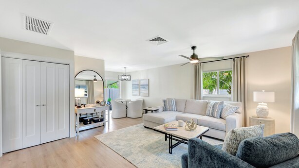 A modern living room with a sofa, coffee table, and armchairs. There is a white wardrobe, a dining area with chairs, and a ceiling fan.