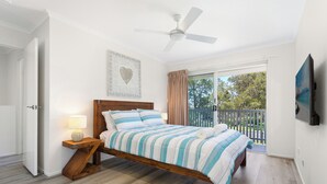 Un dormitorio con cama de madera, una mesita de noche con lámpara, un ventilador de techo y un balcón con vistas a árboles.