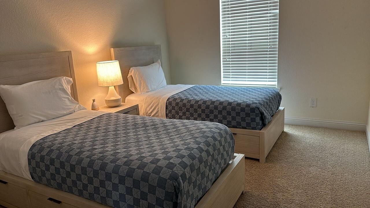 A bedroom with two beds, each with a checkered bedspread, a bedside lamp, and a window with blinds.