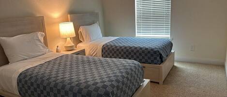 A bedroom with two beds, each with a checkered bedspread, a bedside lamp, and a window with blinds.