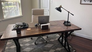 A wooden desk with a laptop, a lamp, and a chair in a room with a patterned rug, a door, and a framed picture on the wall. - Woodlands House. 8 People, Restaurants, Movie Theater, HEB, Parks and Walkways. (Tomball)