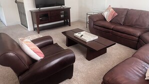 A living room with a brown leather sofa, a matching armchair, a coffee table, a TV on a wooden stand, and a small side table. - Woodlands House. 8 People, Restaurants, Movie Theater, HEB, Parks and Walkways. (Tomball)
