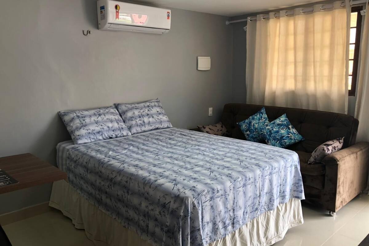 A bedroom with a bed, a sofa, a small table, an air conditioner, and a window with curtains.