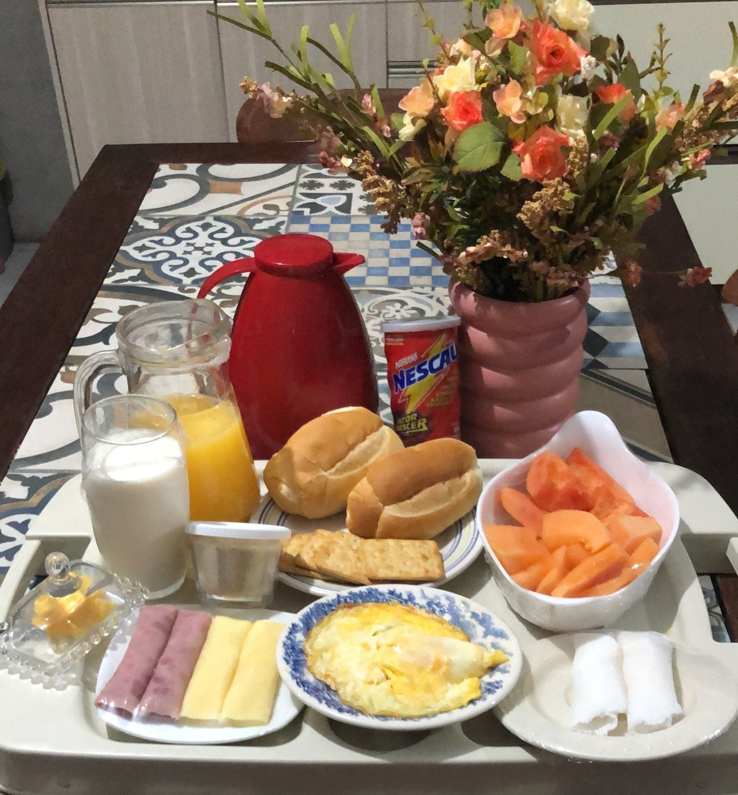 Un plateau de petit-déjeuner comprenant du lait, du jus d’orange, du pain, du jambon, du fromage, des œufs et un arrangement floral.