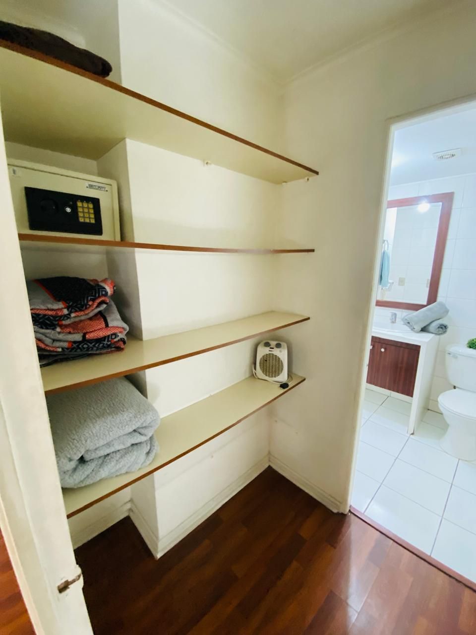 Una esquina de una habitación con estantes de madera, un radiador blanco y una vista parcial de un baño con inodoro y espejo.