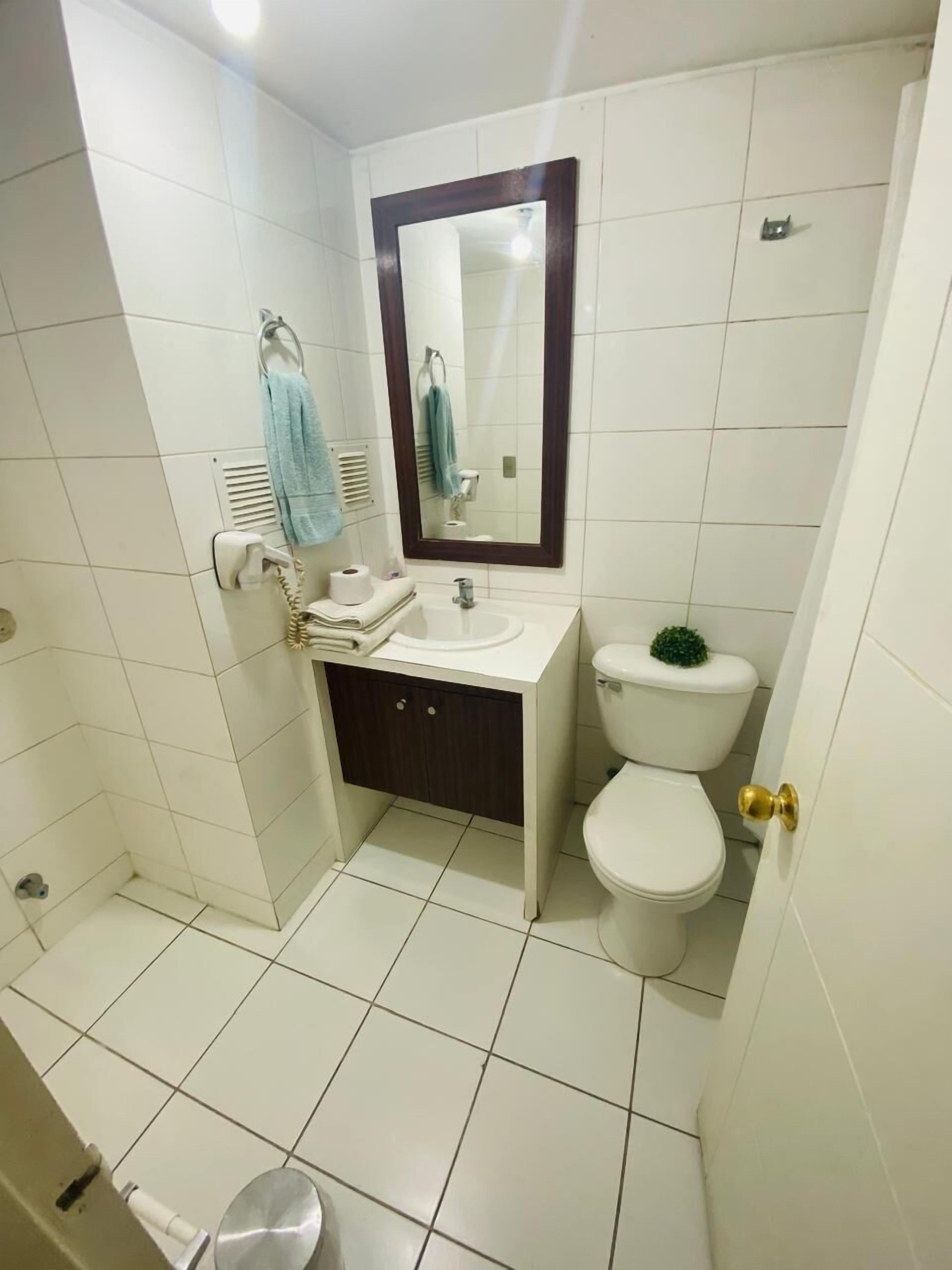A small bathroom with white tiles, a white sink, a mirror, a toilet, and a shower area.