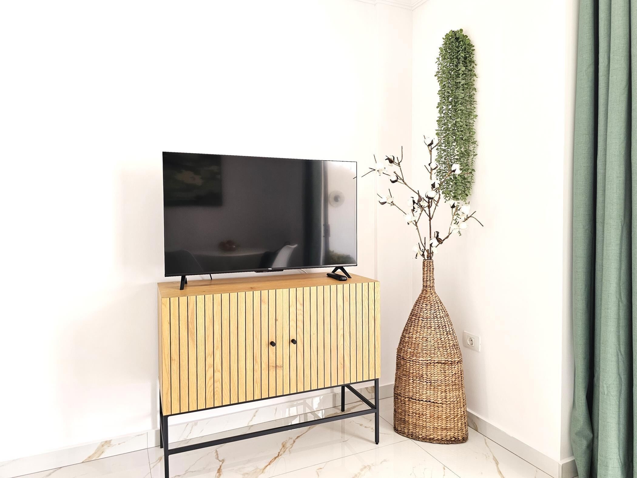 En moderne stue med et fladskærms-tv på et træmøbel, en vævet vase med grene og en grøn gardin.