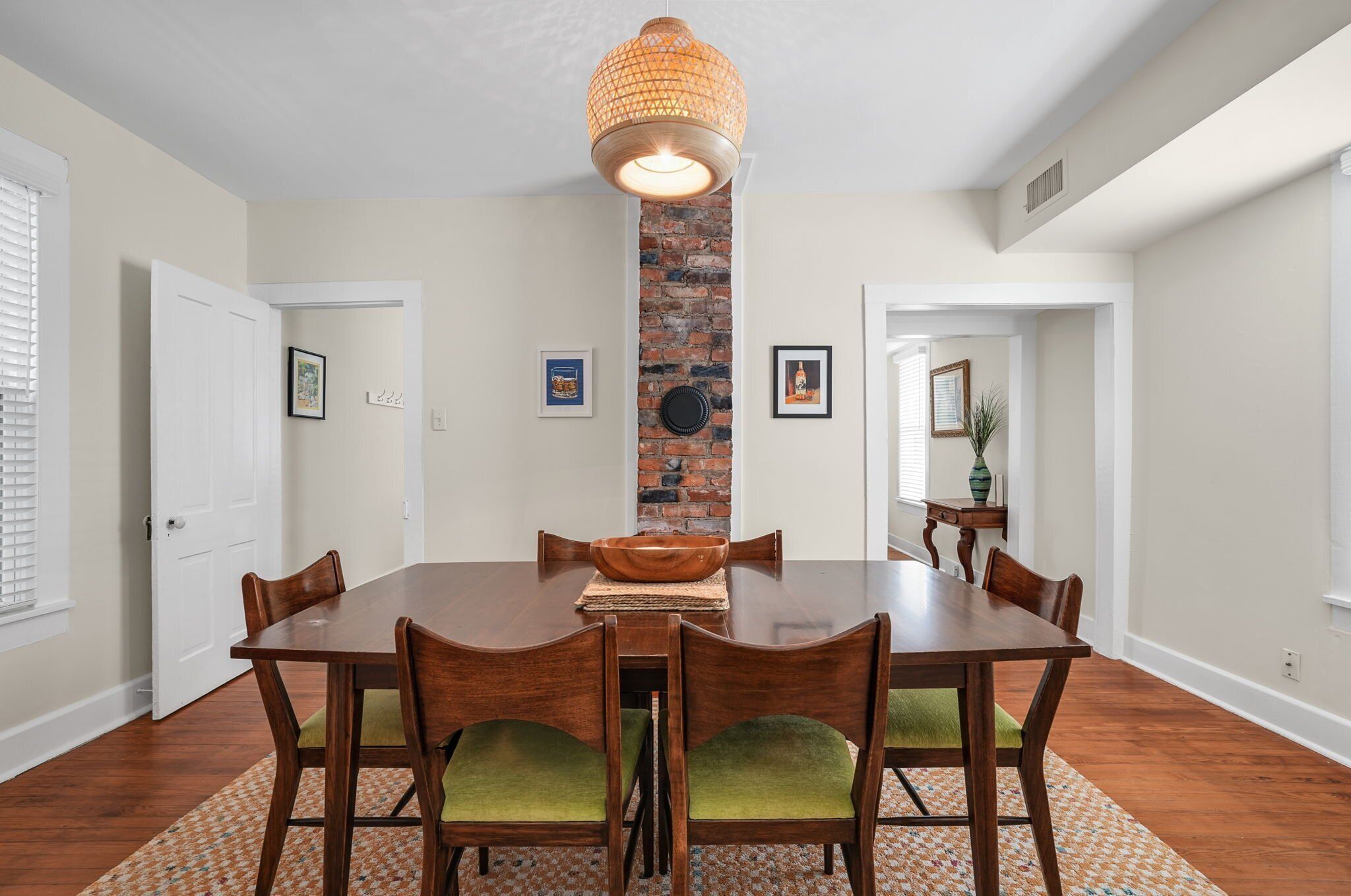 A dining room with a wooden table, green upholstered chairs, a woven light fixture, and a brick accent wall.