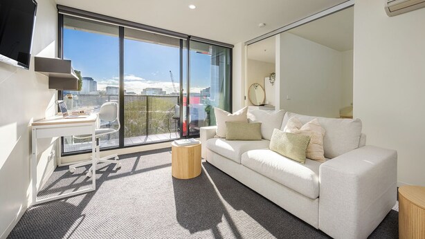A modern living room with a large sliding glass door, a white sofa, a small wooden side table, and a white desk with a chair.
