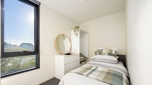A bedroom with a large window, a bedside table with a mirror, a bed with a checkered blanket, and a closet with a hanging plant.