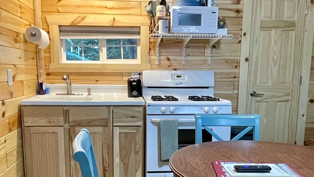 A compact kitchen with wooden walls, a white stove, a microwave, and a small dining area with two chairs. - 
Cabin #3-New, Affordable, Year Round Lodging near Pictured Rocks & Tahquamenon! (Seney)