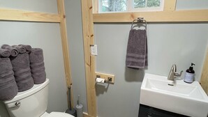 A compact bathroom with a toilet, sink, and window. - 
Cabin #3-New, Affordable, Year Round Lodging near Pictured Rocks & Tahquamenon! (Seney)