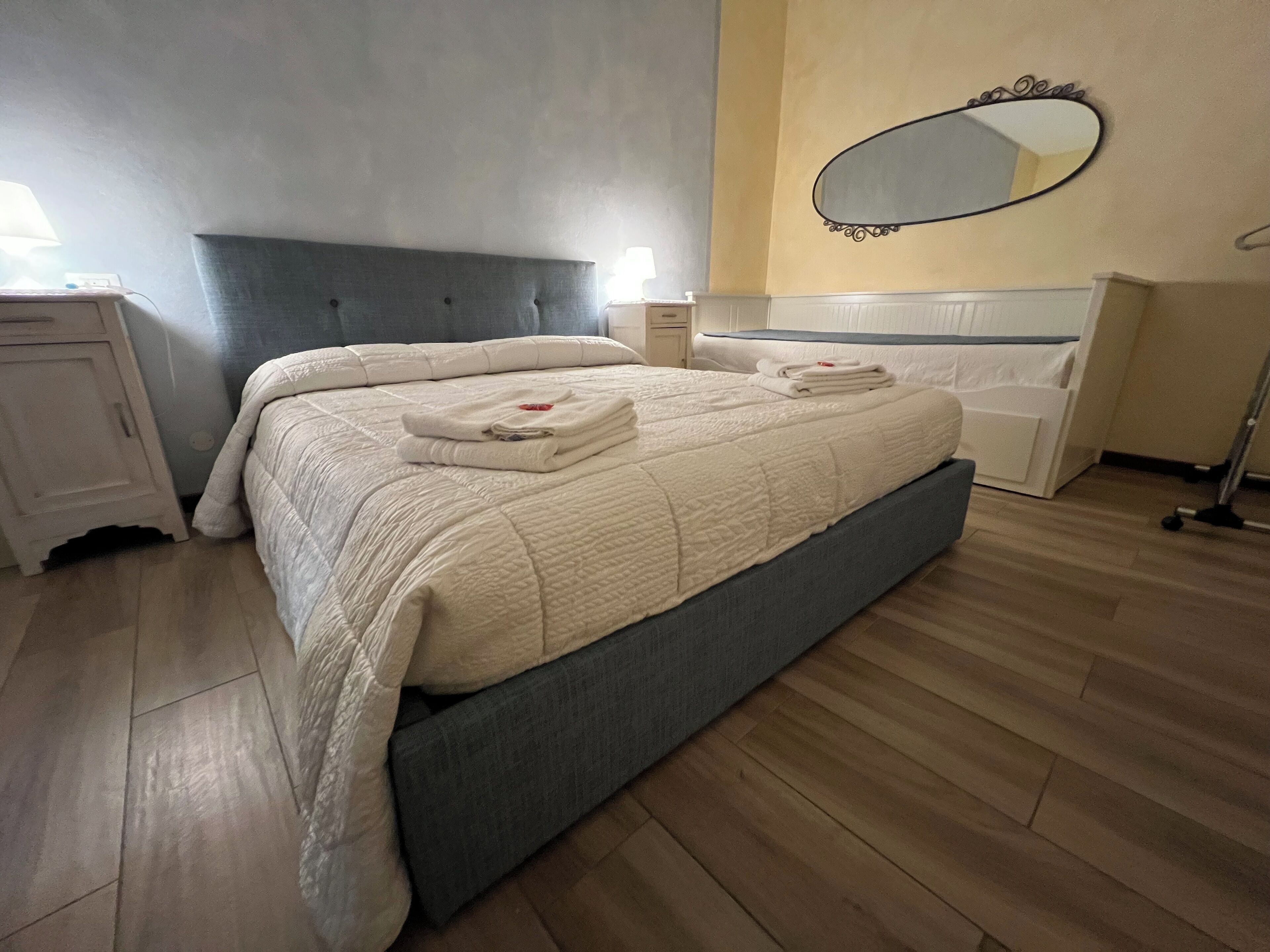 Ein ordentlich bezogenes Bett mit einer Tagesdecke, ein Nachttisch mit Lampe, eine Kommode und ein Spiegel an der Wand.