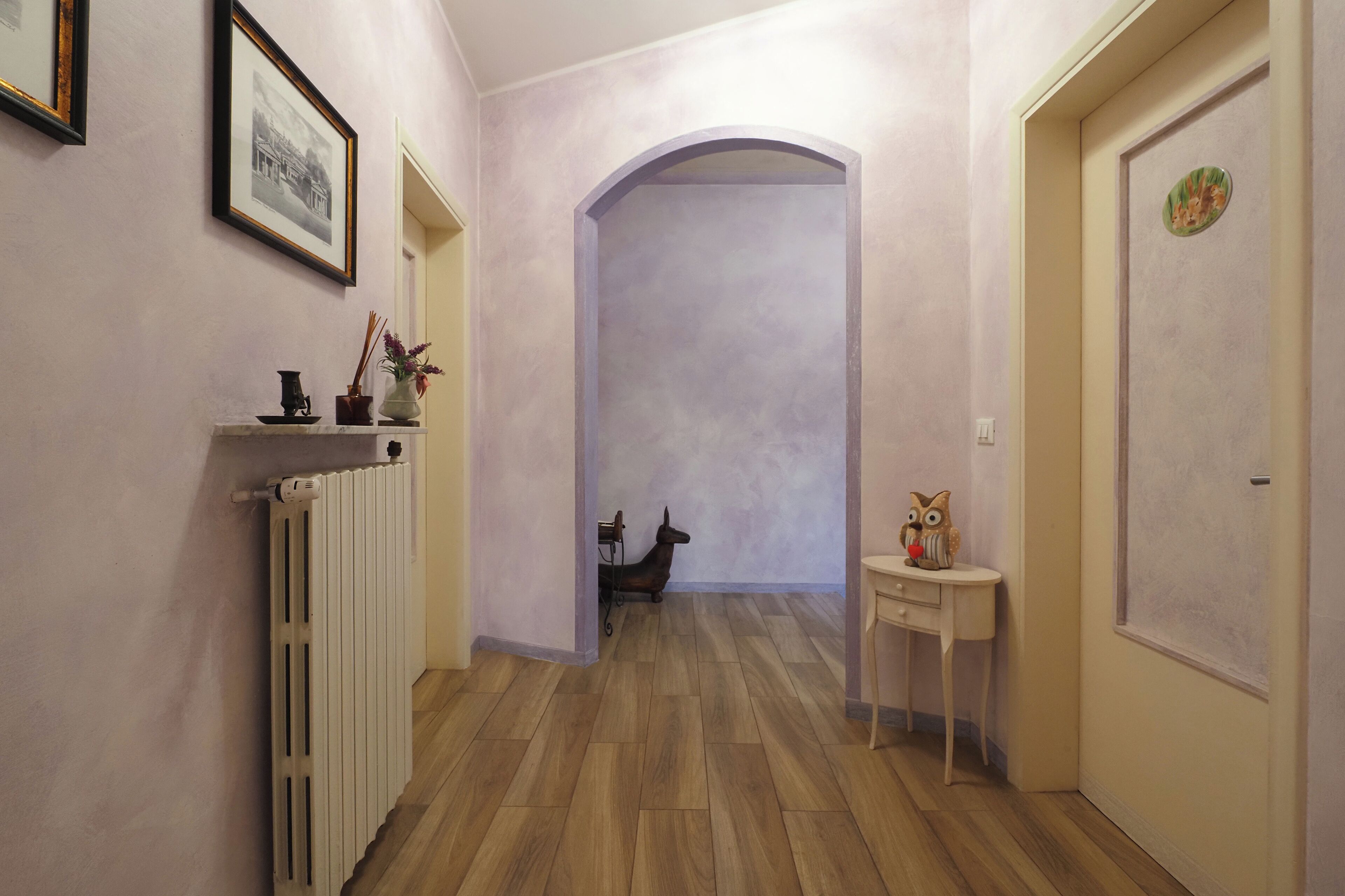 Ein Flur mit Holzboden, einem Heizkörper, einem gerahmten Bild, einem kleinen Tisch mit einer dekorativen Eule und einer Tür mit kreisförmigem Muster.
