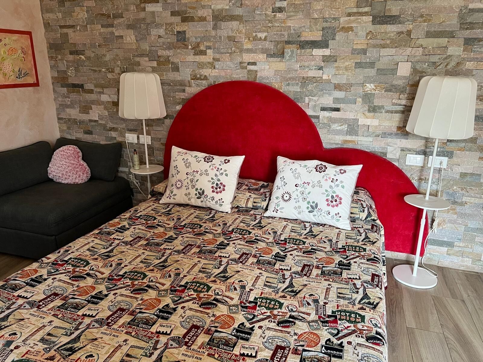 Camera d'albergo con un letto, una coperta con decorazioni a tema viaggio vintage, una testata imbottita rossa e due lampade da comodino.