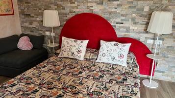 Camera d'albergo con un letto, una coperta con decorazioni a tema viaggio vintage, una testata imbottita rossa e due lampade da comodino.