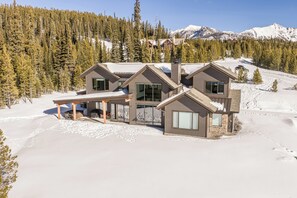 Une maison moderne à plusieurs étages, dotée de grandes fenêtres et d’un patio couvert, entourée d’un paysage enneigé et d’une forêt dense.