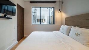 Un dormitorio con una cama grande, un televisor montado en la pared y una ventana con vistas a edificios.