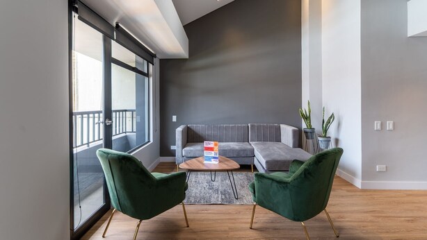 Uma sala de estar moderna com um sofá cinzento, duas poltronas verdes, uma mesa de centro de madeira e uma janela grande com varanda.