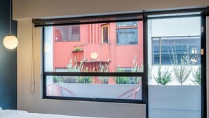 Camera da letto moderna con una grande finestra, un letto e vista su un edificio rosso e del verde circostante.