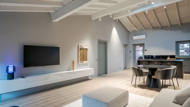 A modern living room with a flat-screen TV, a dining area with a round table and chairs, and a kitchen area with a countertop and appliances. - Attico 56 (Brescia)
