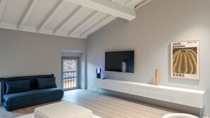 A modern living room with a dark sofa, a white ottoman, a flat-screen TV mounted on a white wall, and a framed poster. - Attico 56 (Brescia)
