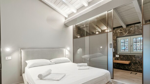 Quarto moderno com uma cama grande, divisória de vidro e vista de um edifício tradicional pela janela.