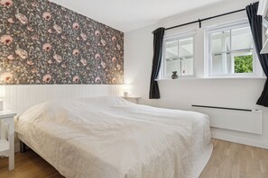 A bedroom with a floral wallpaper, a bed with a white bedspread, a window with curtains, and a bedside table with a lamp. - Renovated summer house in Vejby/ Tisvilde (Vejby)