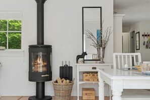 A room with a black wood-burning stove, a white table with chairs, a mirror, and a basket of firewood.
