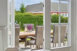 Eine offene Tür führt in einen Außenbereich mit Essplatz, ausgestattet mit Korbstühlen und einem Holztisch. Ein Strauß violetter Blumen steht dort, und man sieht ein rotes Gebäude sowie grüne Pflanzen.