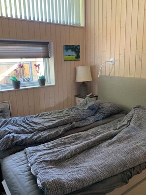 A bedroom with a bed, a window with blinds, a nightstand with a lamp, and a painting on the wall. - Front-row summerhouse with view and big garden (Holbæk)