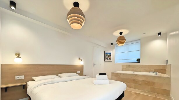 A modern hotel room with a large bed, bedside lamps, a built-in vanity with a bathtub, and a large window. - Les Fauvettes (Épernay)