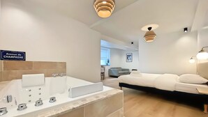 A modern hotel room with a large bathtub, a bed, and a dining area in the background. - Les Fauvettes (Épernay)