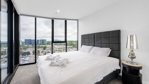 Une chambre d’hôtel moderne avec un grand lit, une table de chevet et une vue sur le paysage urbain grâce à de grandes fenêtres du sol au plafond.