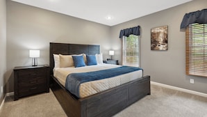 A bedroom with a dark wooden bed frame, a beige quilt, and blue pillows. There are two bedside tables with lamps, a window with blinds, and a wall-mounted picture. - Hatton Hideaway at Red River Gorge! Pool / Hot Tub / Arcade Game Room (Stanton)