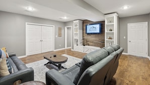 Un salon moderne avec un canapé en cuir, une table basse et un téléviseur fixé à un mur en bois. On y trouve des portes blanches et des étagères intégrées ornées d’objets décoratifs.