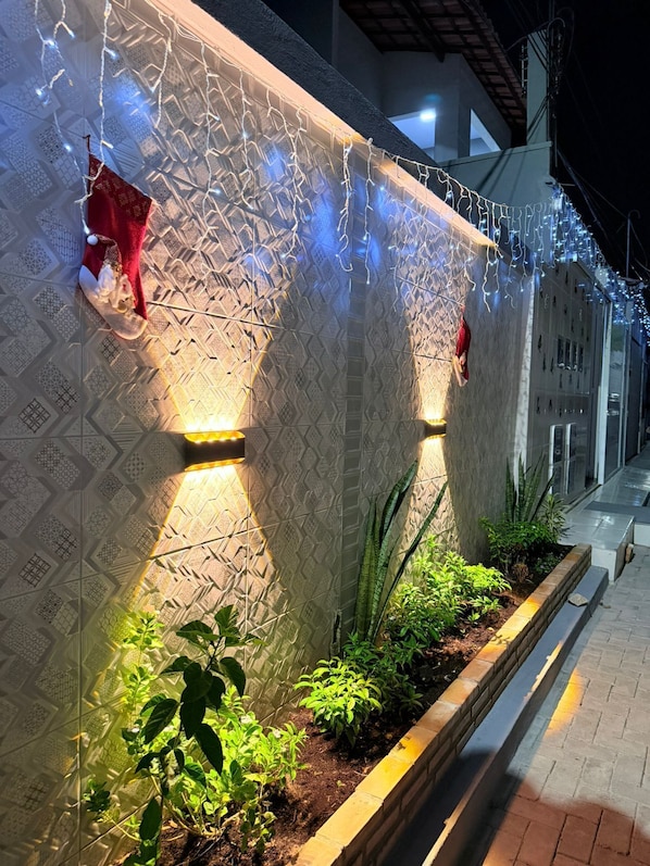 A building facade with patterned wall design, illuminated by warm lights, and bordered by a planter with green plants.