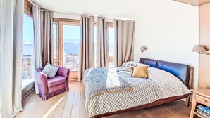 A bedroom with a bed, a chair, a nightstand, and a view of mountains through the window. - Jeanne D'Arc LOFT Lac by Interhome (Crans-Montana)