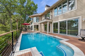 A swimming pool with a wooden deck, a covered patio with outdoor furniture, and a large glass door leading to a covered balcony.
